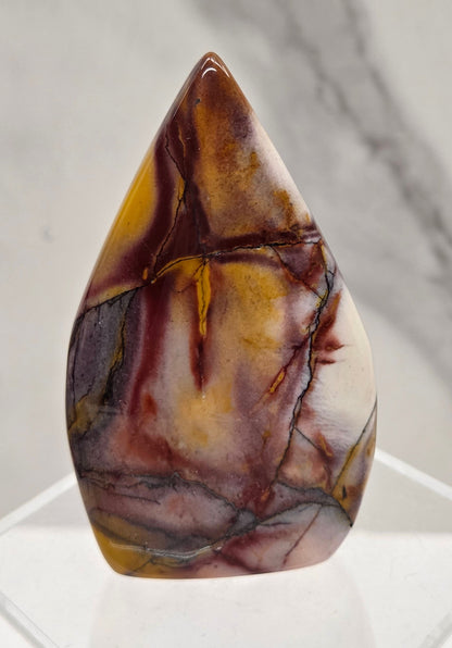 A polished teardrop-shaped cabochon gemstone with marbled brown, gold, and cream patterns, displayed on a clear acrylic stand against a light gray marble background.
