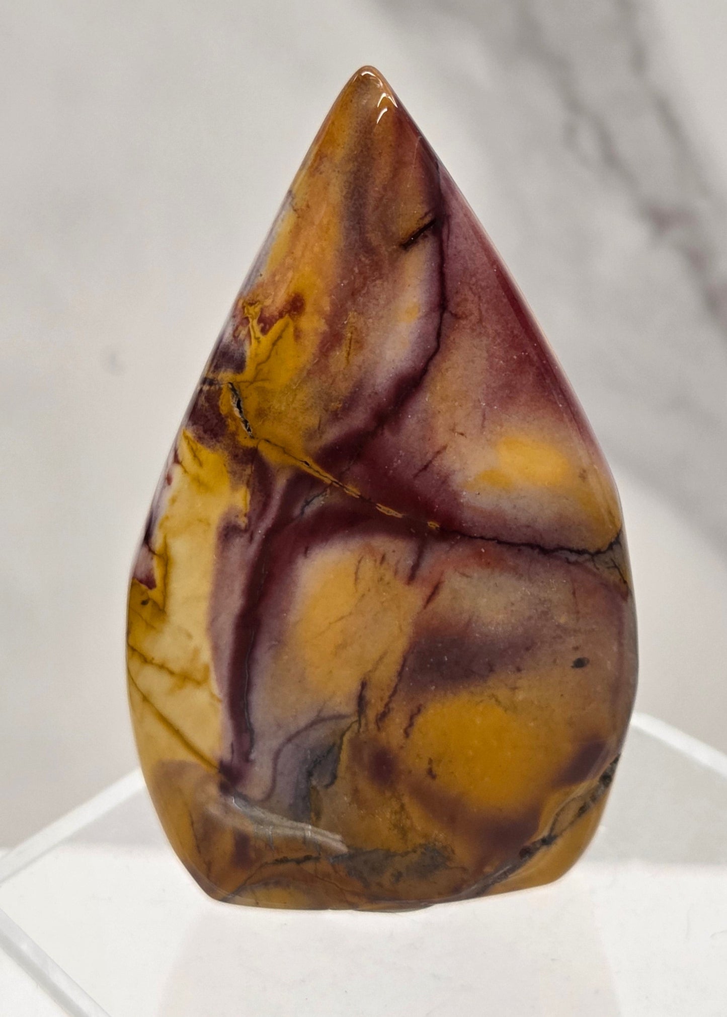 A polished teardrop-shaped gemstone cabochon with marbled patterns of golden yellow, brown, and deep purple, displayed on a clear acrylic stand against a light gray marble background.