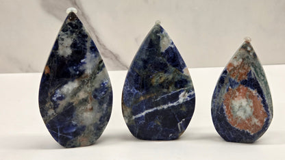 Three teardrop-shaped polished gemstone pendants in shades of lapis lazuli, set against a white marble-patterned background.