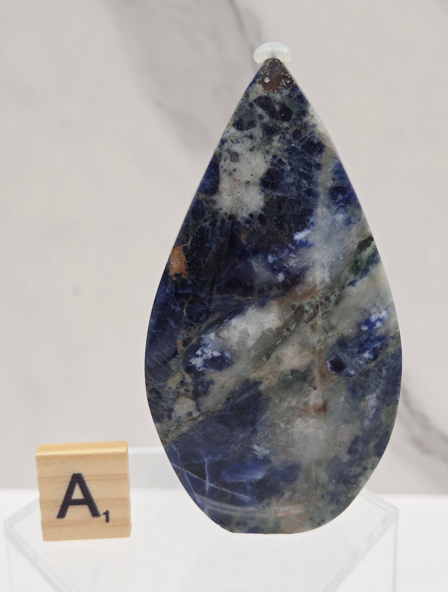A polished teardrop-shaped gemstone with a marbled pattern of deep blue, white, and gray, displayed on a clear acrylic stand next to a small wooden letter tile on a white marble surface.