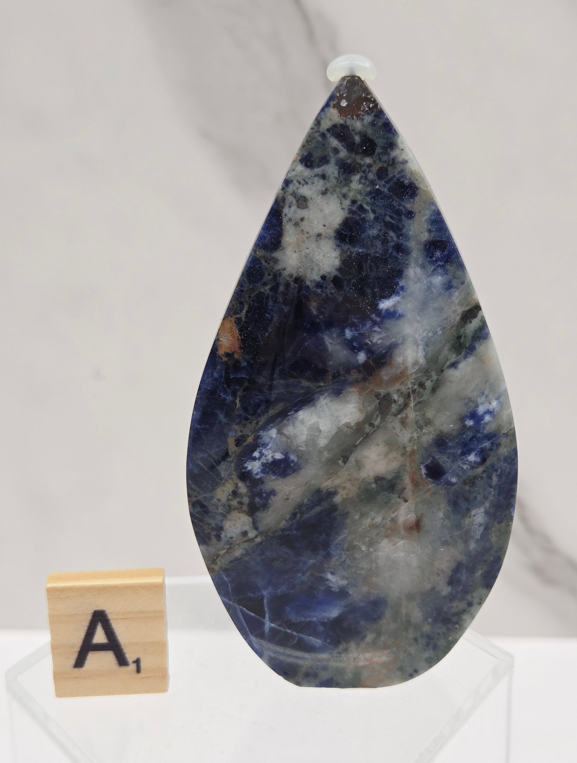 A polished teardrop-shaped gemstone with a marbled pattern of deep blue, white, and gray, displayed on a clear acrylic stand next to a small wooden letter tile on a white marble surface.