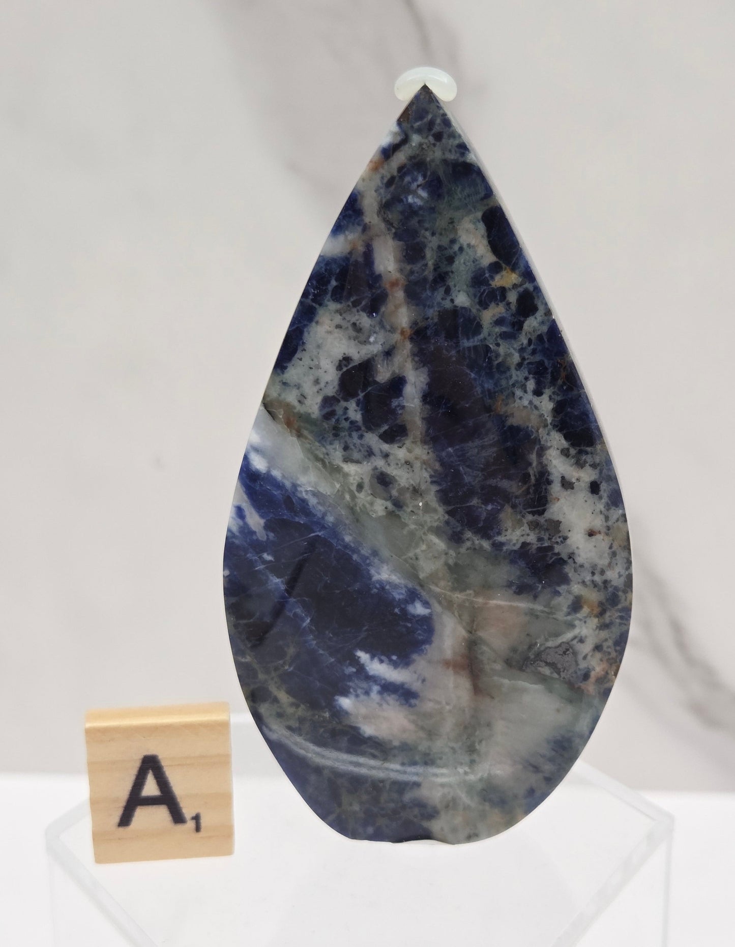 A teardrop-shaped polished stone slab with a marbled pattern of deep blue, white, and gray, displayed on a white marble surface next to a small wooden letter tile.