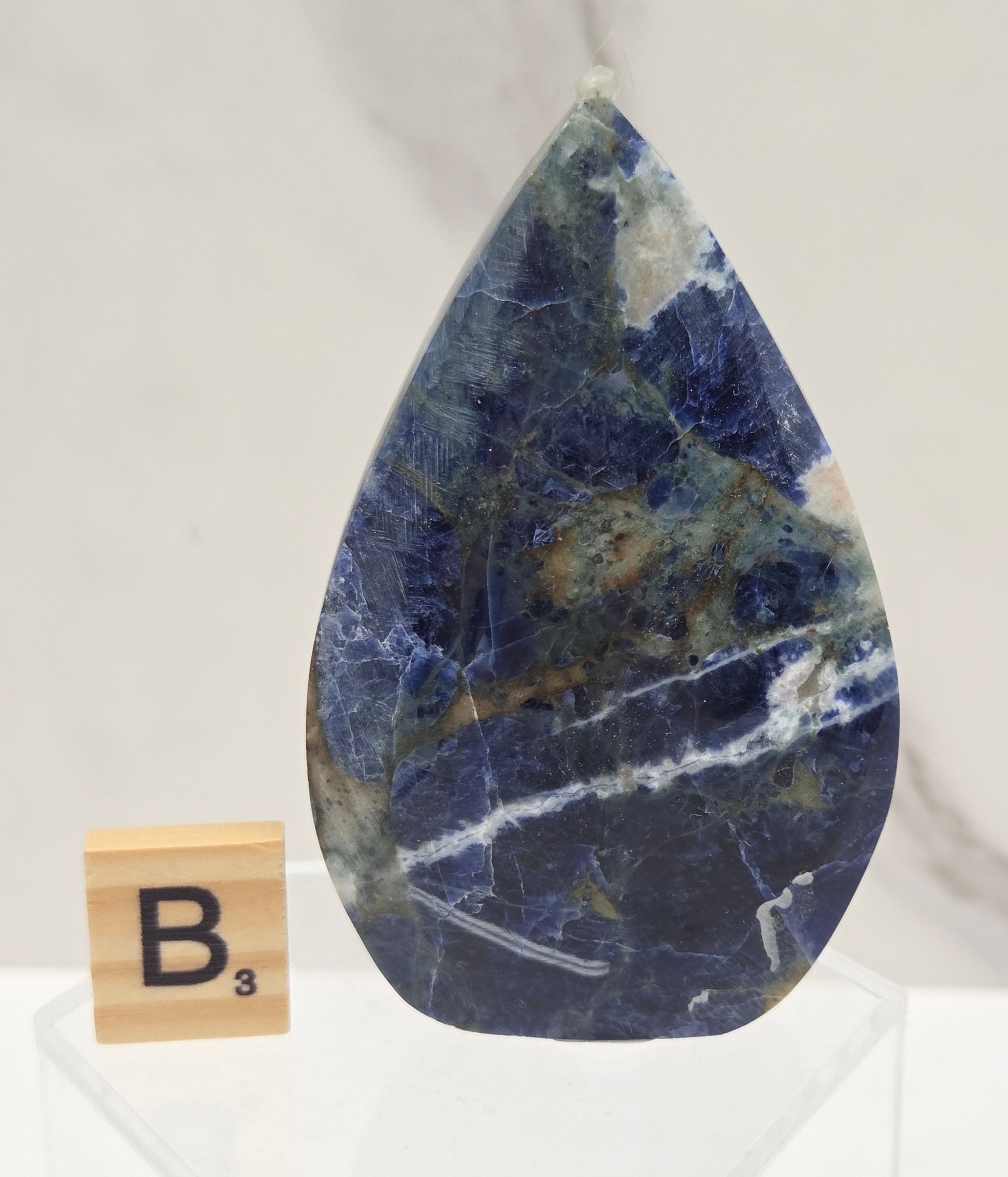 A polished blue stone tile with natural white and gray veining, displayed on a white marble surface next to a small wooden tile labeled B3.