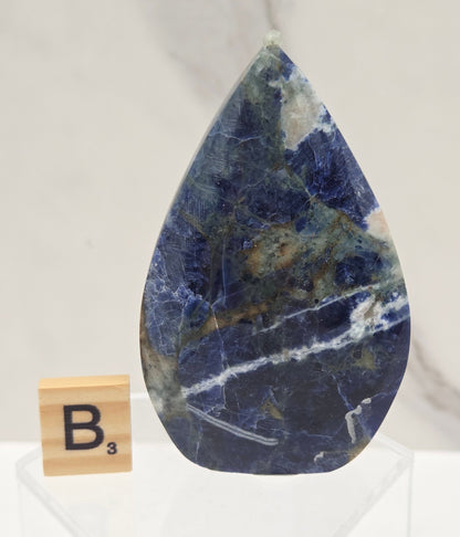 A polished blue stone tile with natural white and gray veining, displayed on a white marble surface next to a small wooden tile labeled B3.