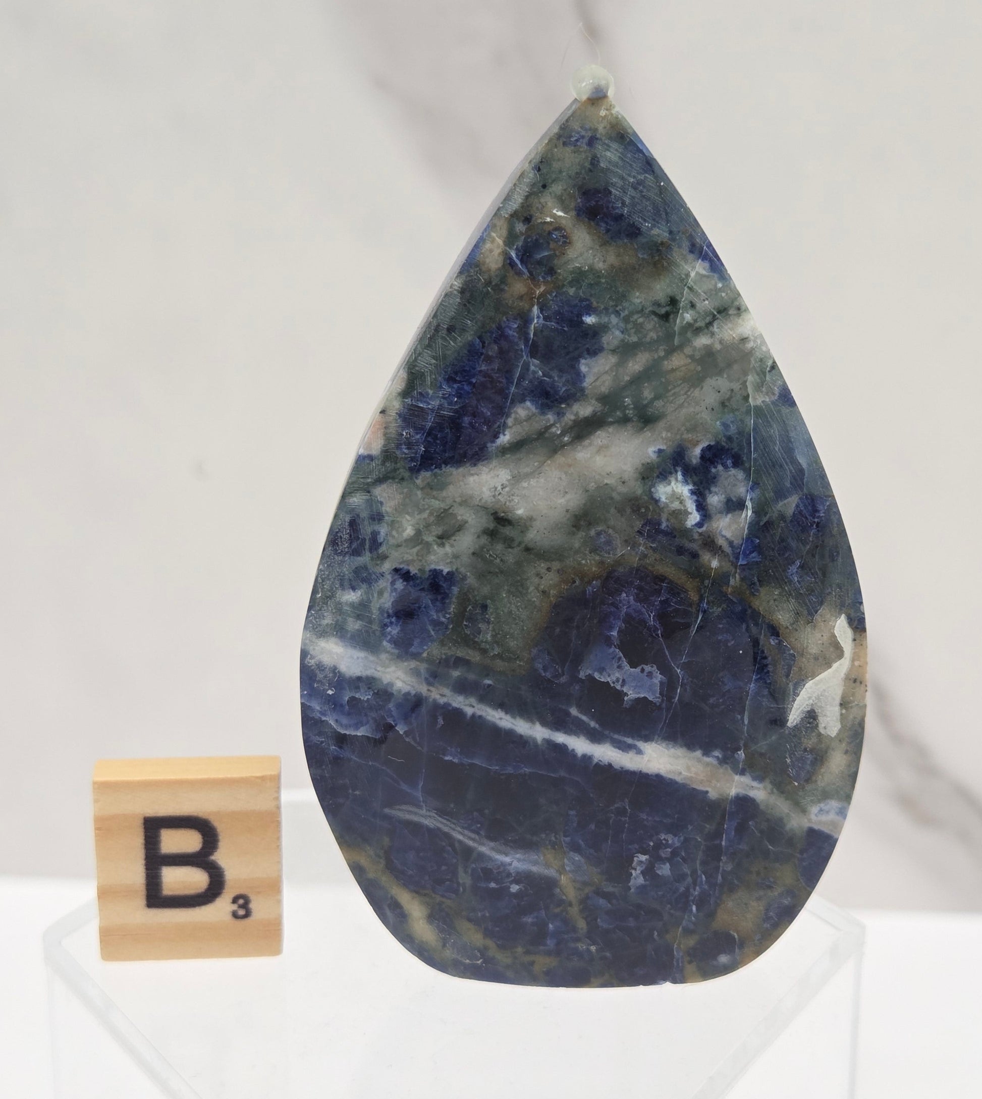 A polished teardrop-shaped blue stone, likely lapis lazuli, displayed on a clear acrylic stand next to a wooden Scrabble tile with the letter 'B'. The background is a white marble surface.