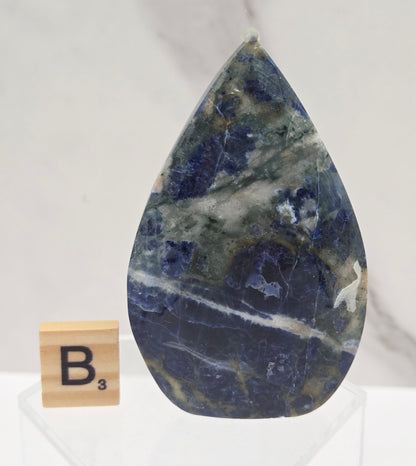 A polished teardrop-shaped blue stone, likely lapis lazuli, displayed on a clear acrylic stand next to a wooden Scrabble tile with the letter 'B'. The background is a white marble surface.