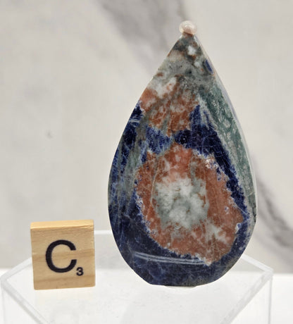 A polished teardrop-shaped lapis lazuli crystal pendant with a natural band of copper and white matrix, displayed on a clear acrylic stand next to a wooden Scrabble tile on a white marble surface.