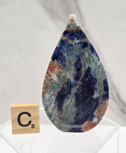 A teardrop-shaped polished gemstone with a marbled pattern of navy blue, green, and brown, displayed on a clear acrylic stand next to a wooden Scrabble tile on a white marble surface.
