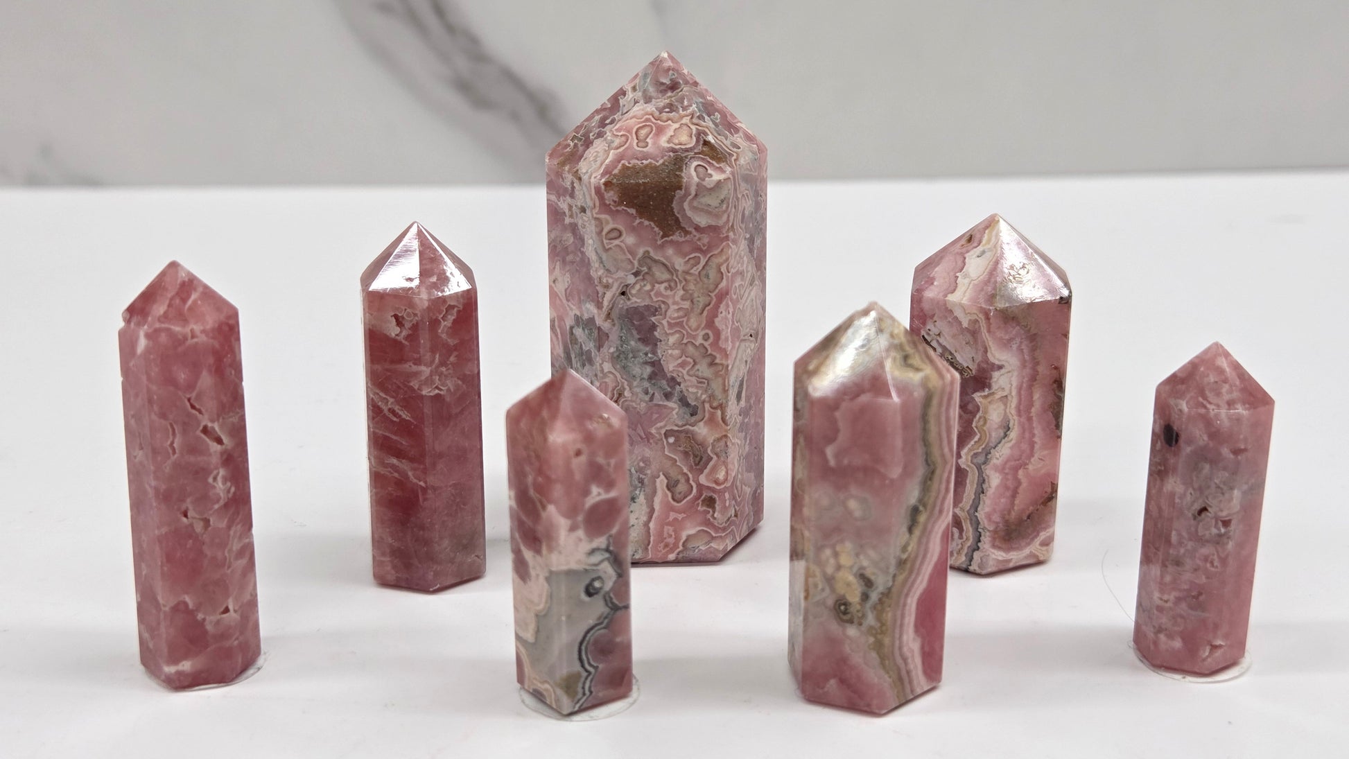 Seven polished pinkish-red stone crystal points of various heights arranged on a white surface with a light gray marble-patterned background.