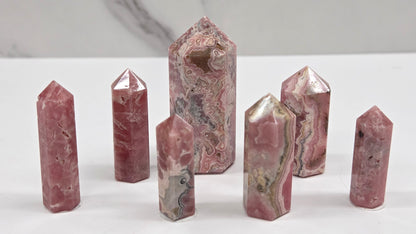 Seven polished pinkish-red stone crystal points of various heights arranged on a white surface with a light gray marble-patterned background.