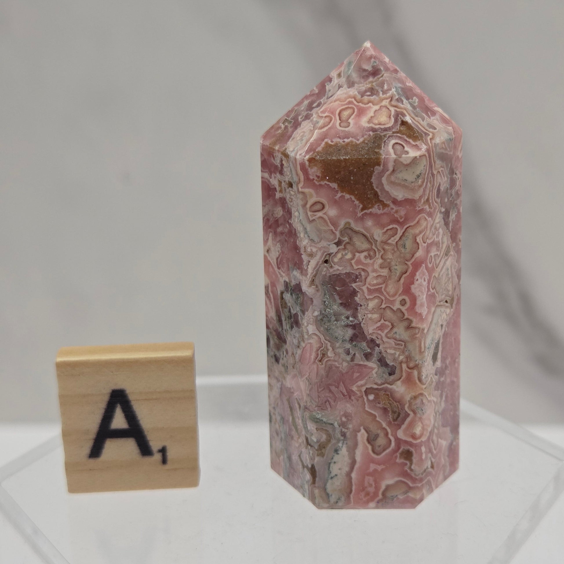 A polished pink and white agate crystal point displayed on a clear acrylic stand, with a wooden letter tile showing the letter 'A' placed below it for scale, against a light gray marble background.
