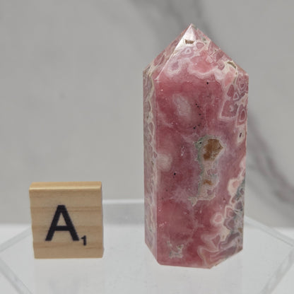 A pink agate crystal point with natural white and gray banded patterns, displayed on a clear acrylic stand next to a wooden letter tile.