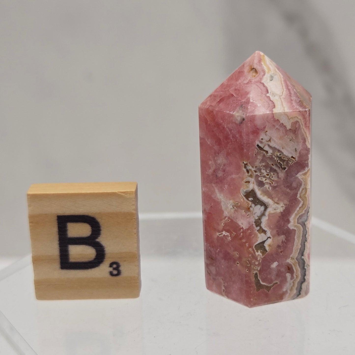 A polished pinkish-red stone point with natural banding and metallic gold inclusions, displayed above a wooden Scrabble tile on a clear acrylic stand against a light marble-like background.