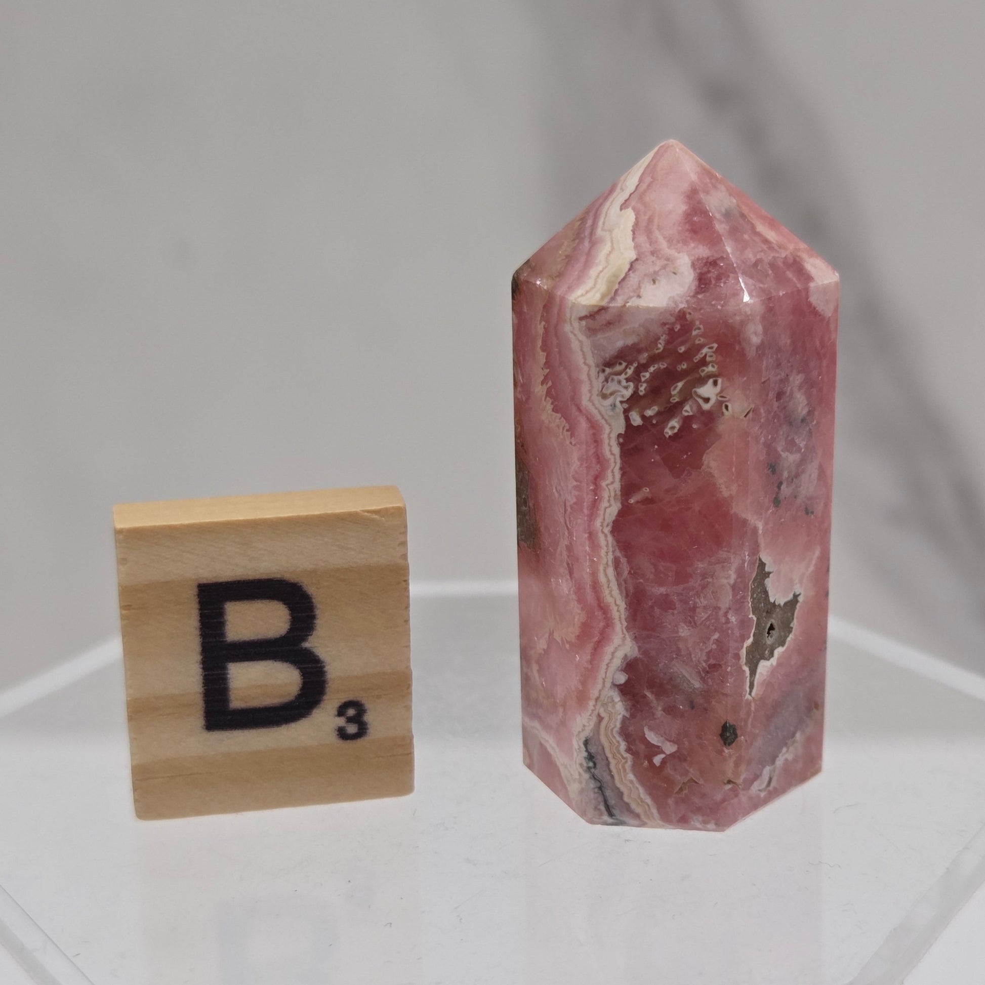 A pinkish-red agate crystal point with natural banding and metallic inclusions, displayed above a wooden Scrabble tile marked 'B3' on a clear acrylic stand against a light gray marble background.