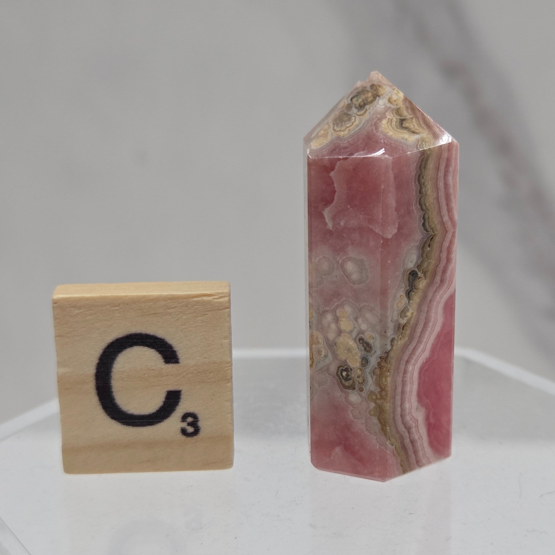 A polished pink and beige banded stone crystal point displayed on a clear acrylic stand above a Scrabble tile showing the letter 'C' and value 3.