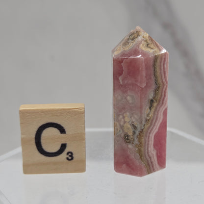 A polished pink and beige banded stone crystal point displayed on a clear acrylic stand above a Scrabble tile showing the letter 'C' and value 3.
