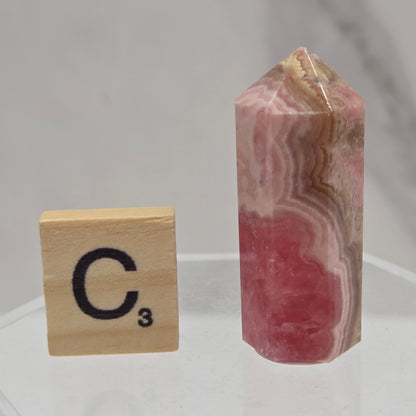 A polished pink and white banded stone cylinder with a pointed end, displayed above a wooden Scrabble tile showing the letter 'C' and number 3, against a light gray marble background.
