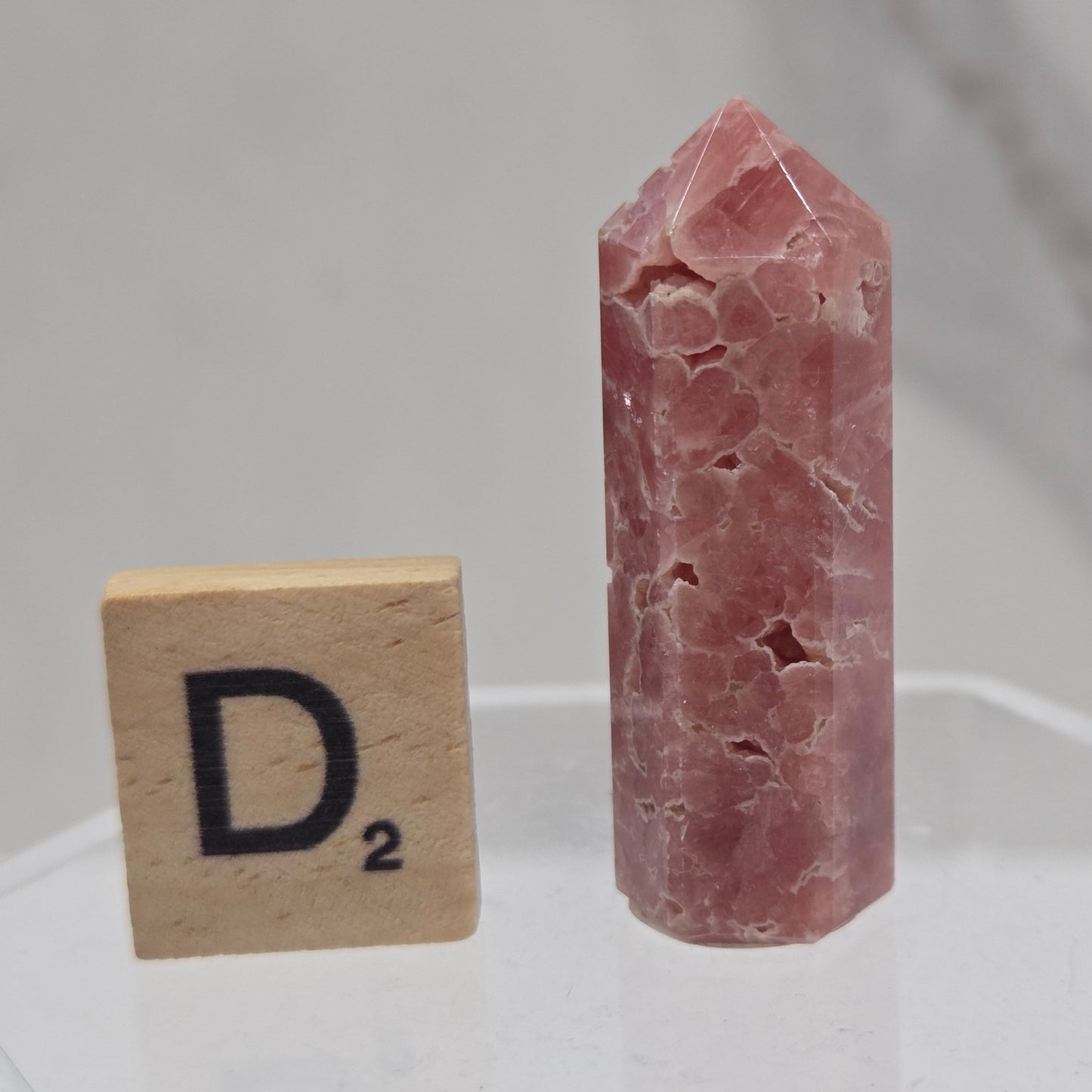 A pinkish-red rose quartz crystal point displayed on a clear acrylic stand, with a wooden D2 tile below for scale, against a light gray marble background.