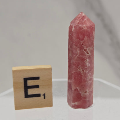 A pink rose quartz crystal point displayed horizontally on a clear acrylic stand, with a small wooden tile showing the letter 'E' below it.