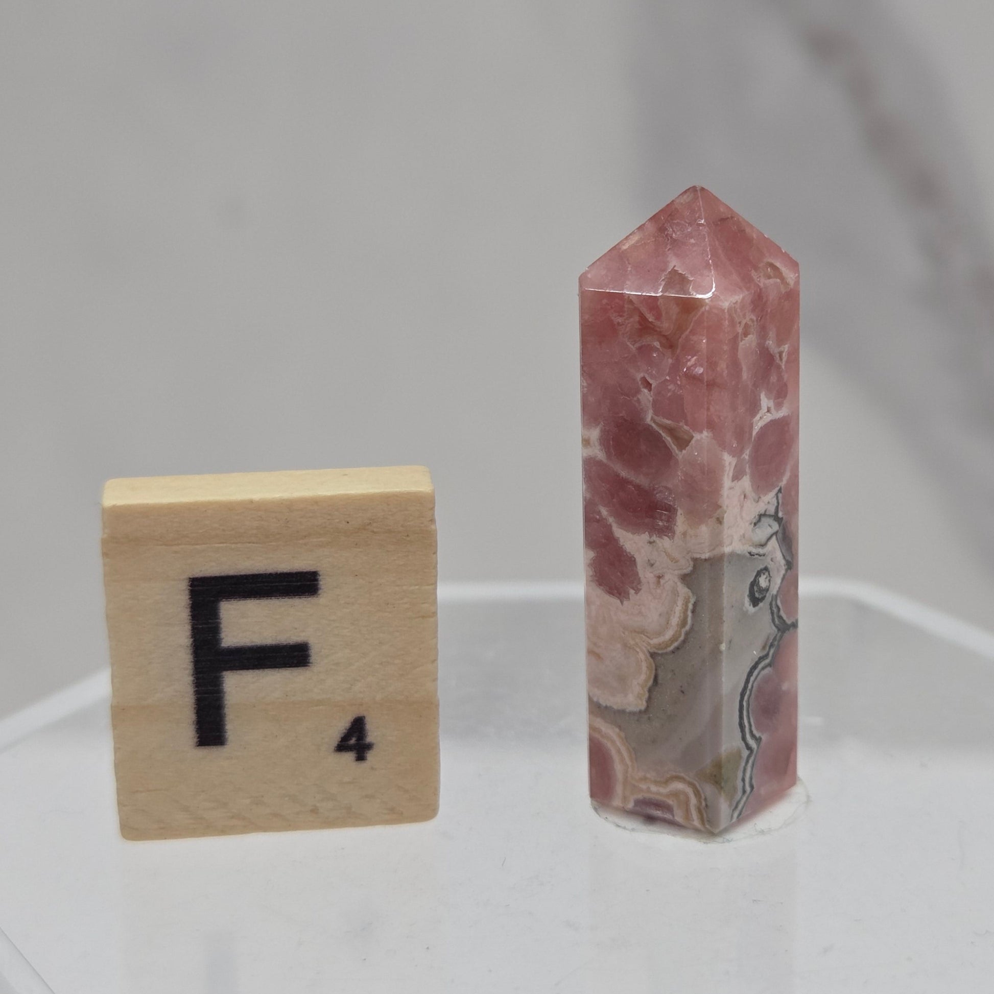 A polished pinkish-red stone point crystal displayed vertically on a clear acrylic stand, with a wooden Scrabble tile showing the letter F placed below it for scale, against a light gray marble-patterned background.