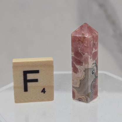 A polished pinkish-red stone point crystal displayed vertically on a clear acrylic stand, with a wooden Scrabble tile showing the letter F placed below it for scale, against a light gray marble-patterned background.