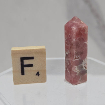 A small polished pinkish-red crystal point displayed horizontally on a clear acrylic stand, with a wooden Scrabble tile showing the letter F placed below it for scale, against a light gray marble-like background.