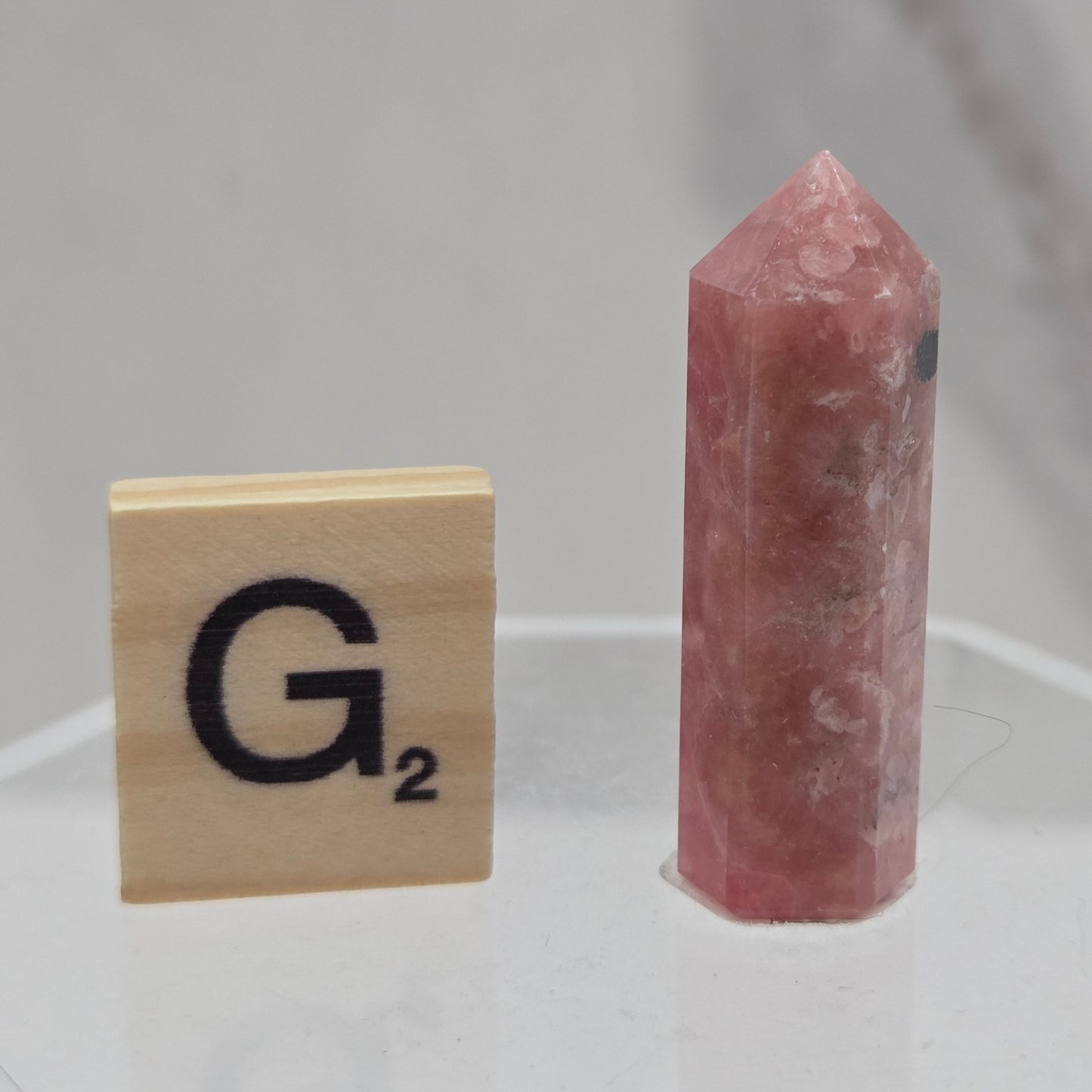 A small pinkish-red crystal point displayed horizontally above a wooden tile with a black 'G2' symbol, set against a light gray marble-patterned background.