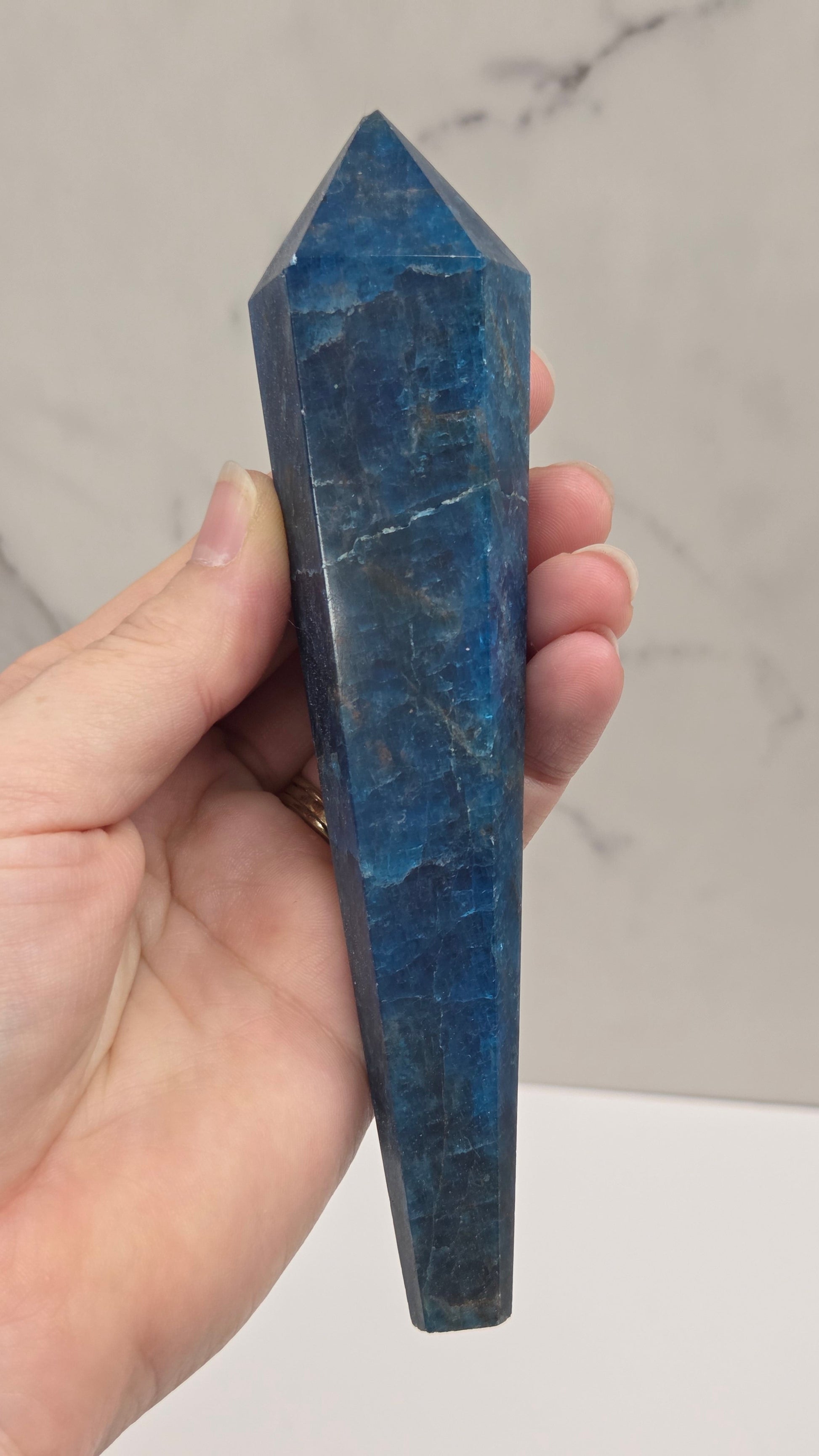 A hand holding a polished blue apatite crystal point against a light marble-patterned background.