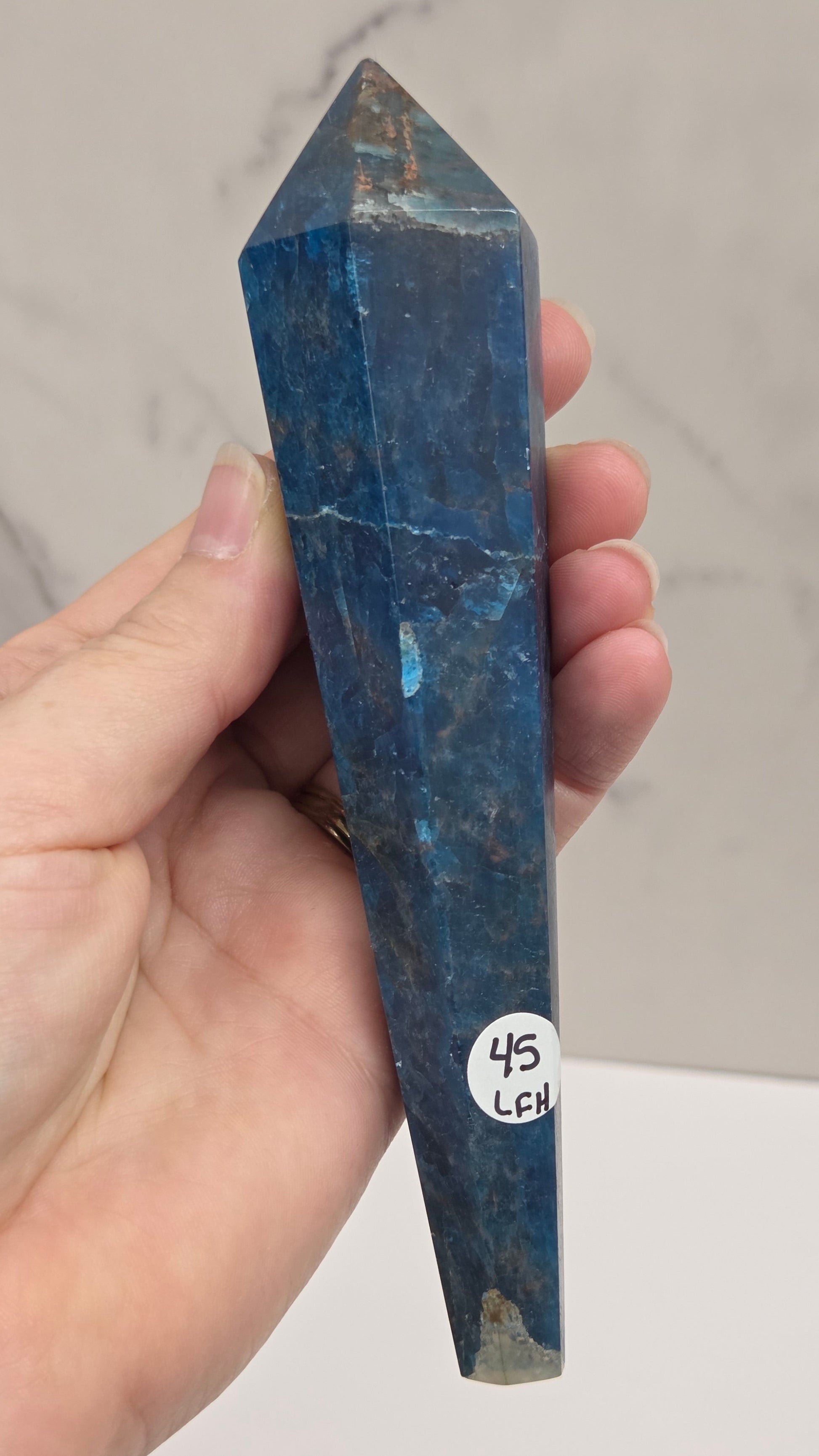 A hand holding a blue apatite crystal point against a light gray marble-patterned background.