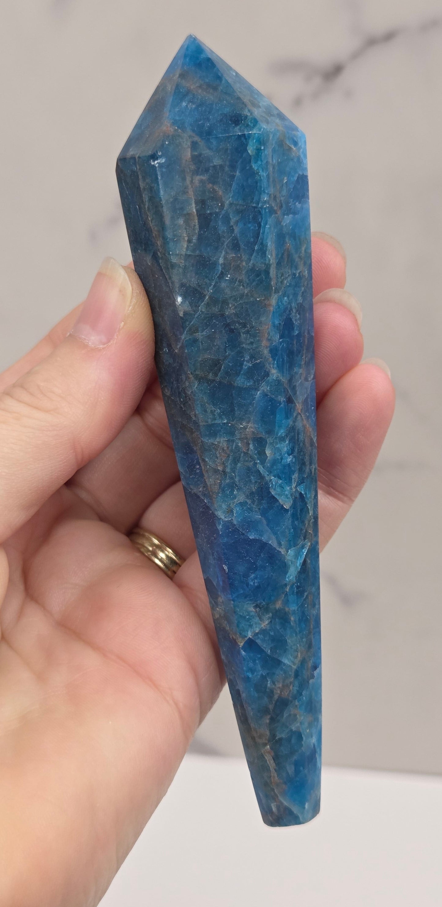 A hand holding a polished blue apatite crystal point against a light marble-patterned background.