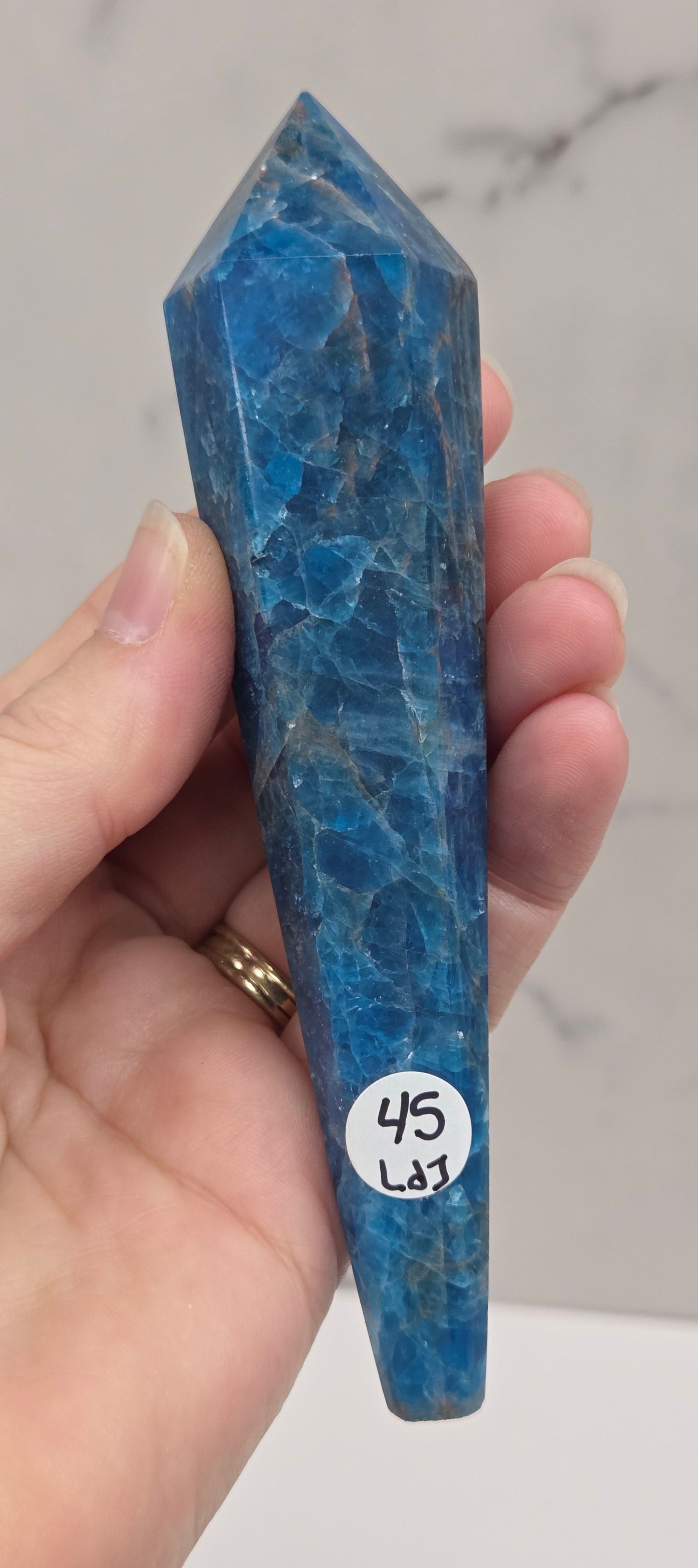A hand holding a polished blue apatite crystal point against a light marble-patterned background. A small white price tag sticker is attached to the crystal.
