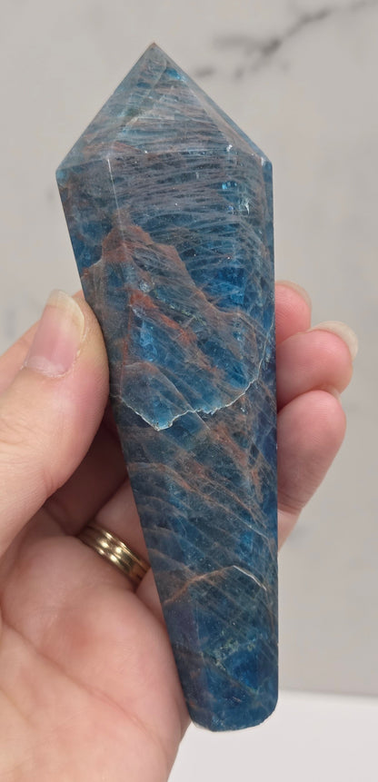 A hand holding a polished blue apatite crystal point against a light marble background.