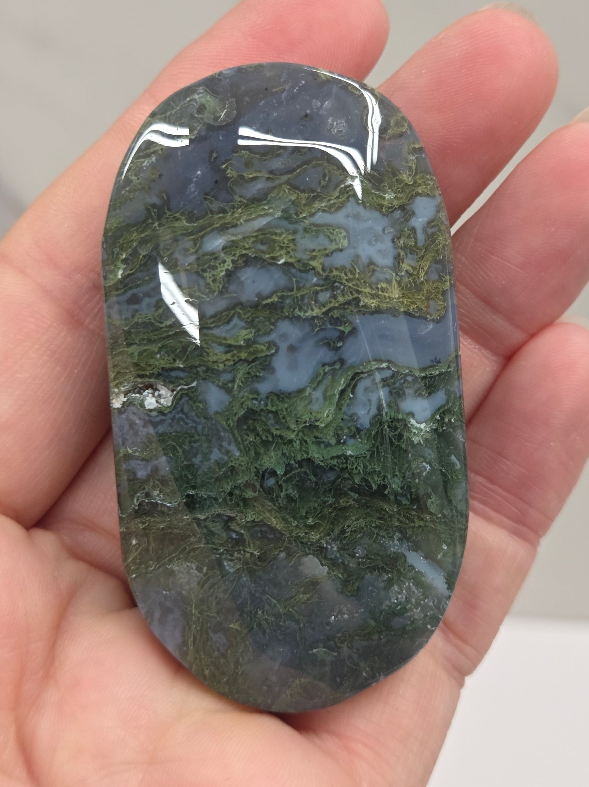 A polished dark green and gray stone with natural marbled patterns, held in a person's hand against a light gray marble background.