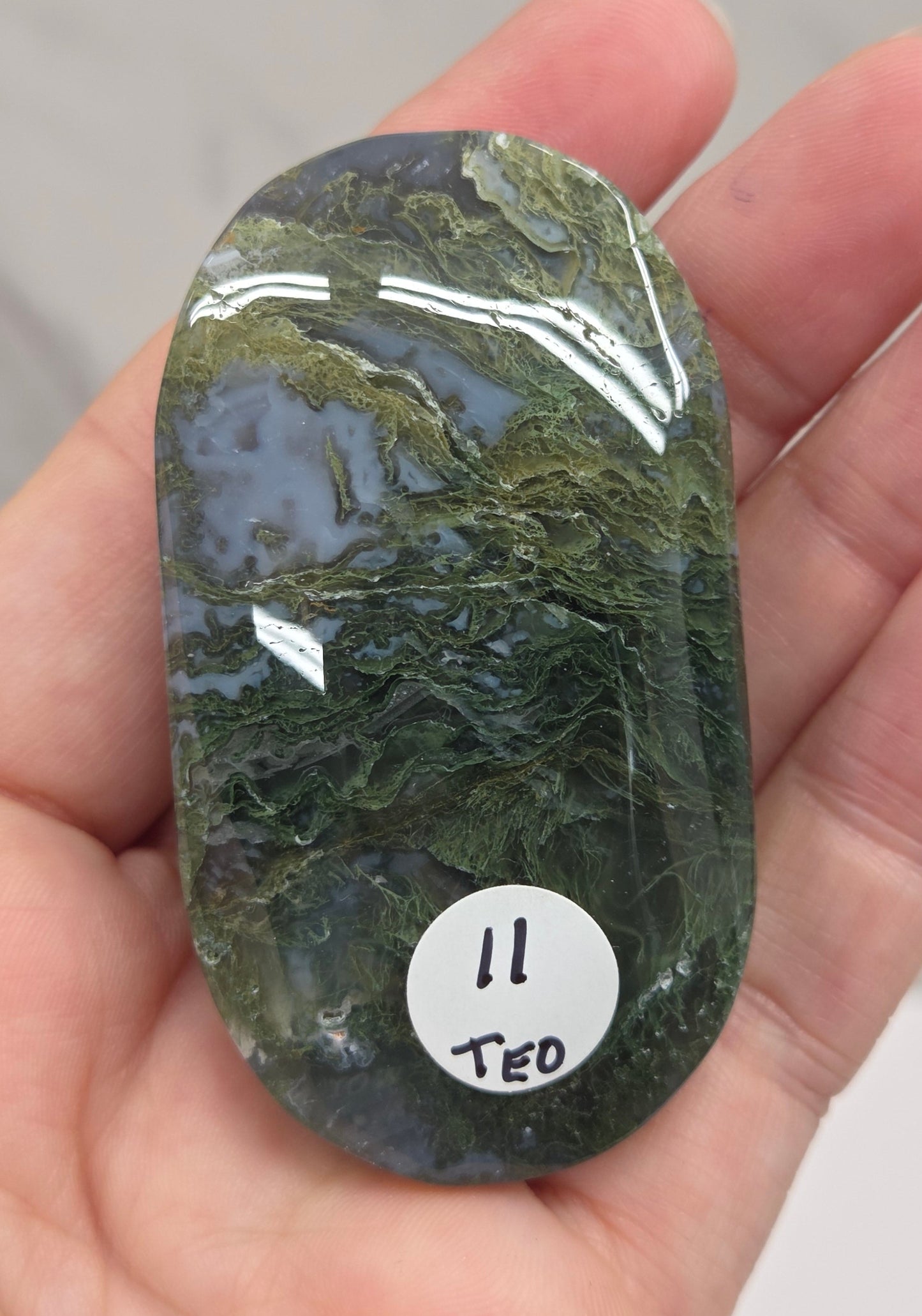 A hand holding a polished green stone or crystal with natural marbled patterns, featuring a small white sticker labeled '11 TEO'.