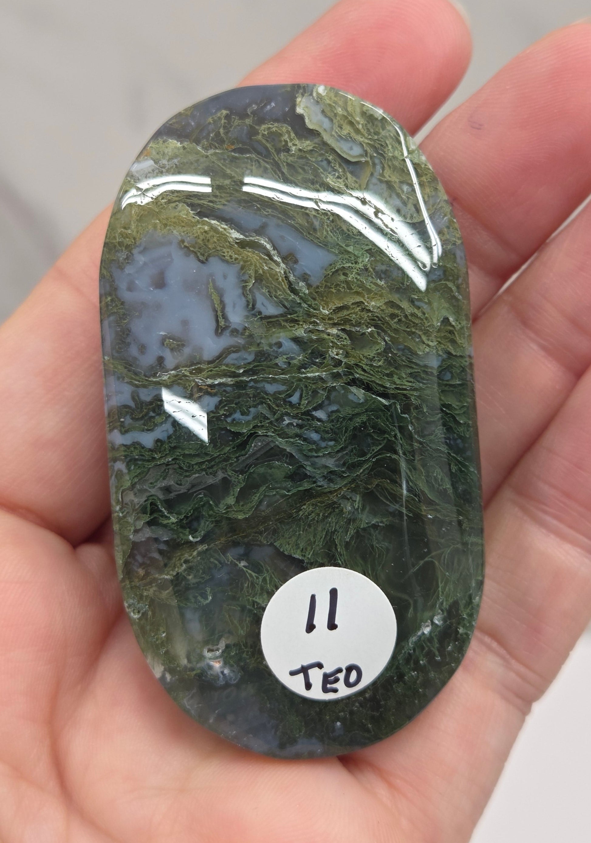 A hand holding a polished green stone or crystal with natural marbled patterns, featuring a small white sticker labeled '11 TEO'.