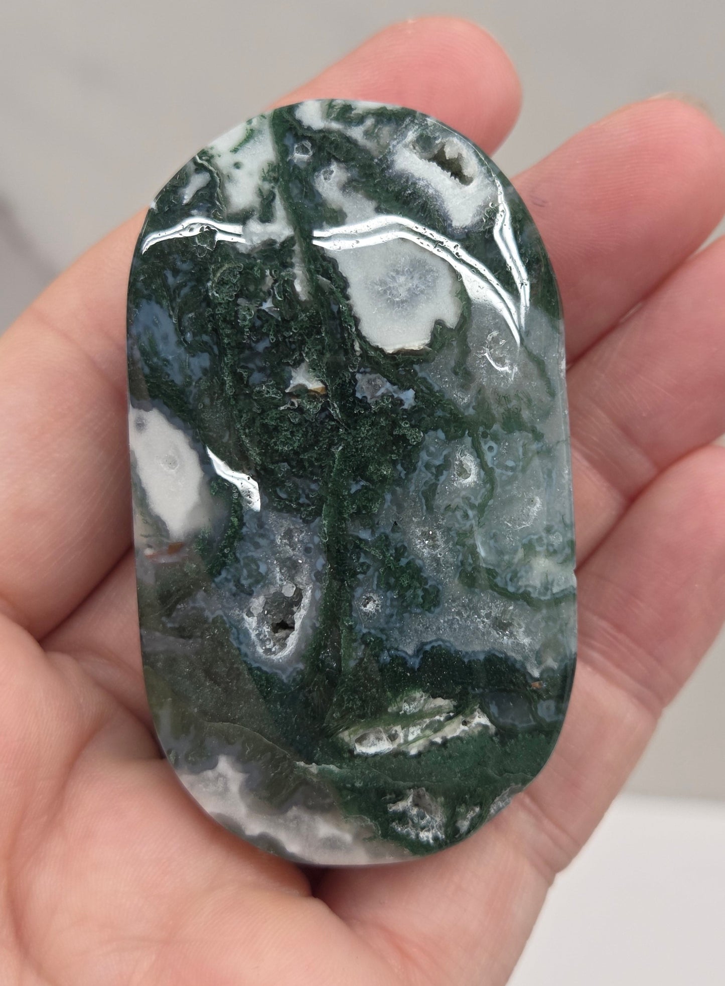 A hand holding a polished dark green and white marbled stone or crystal against a light gray marble-like background.