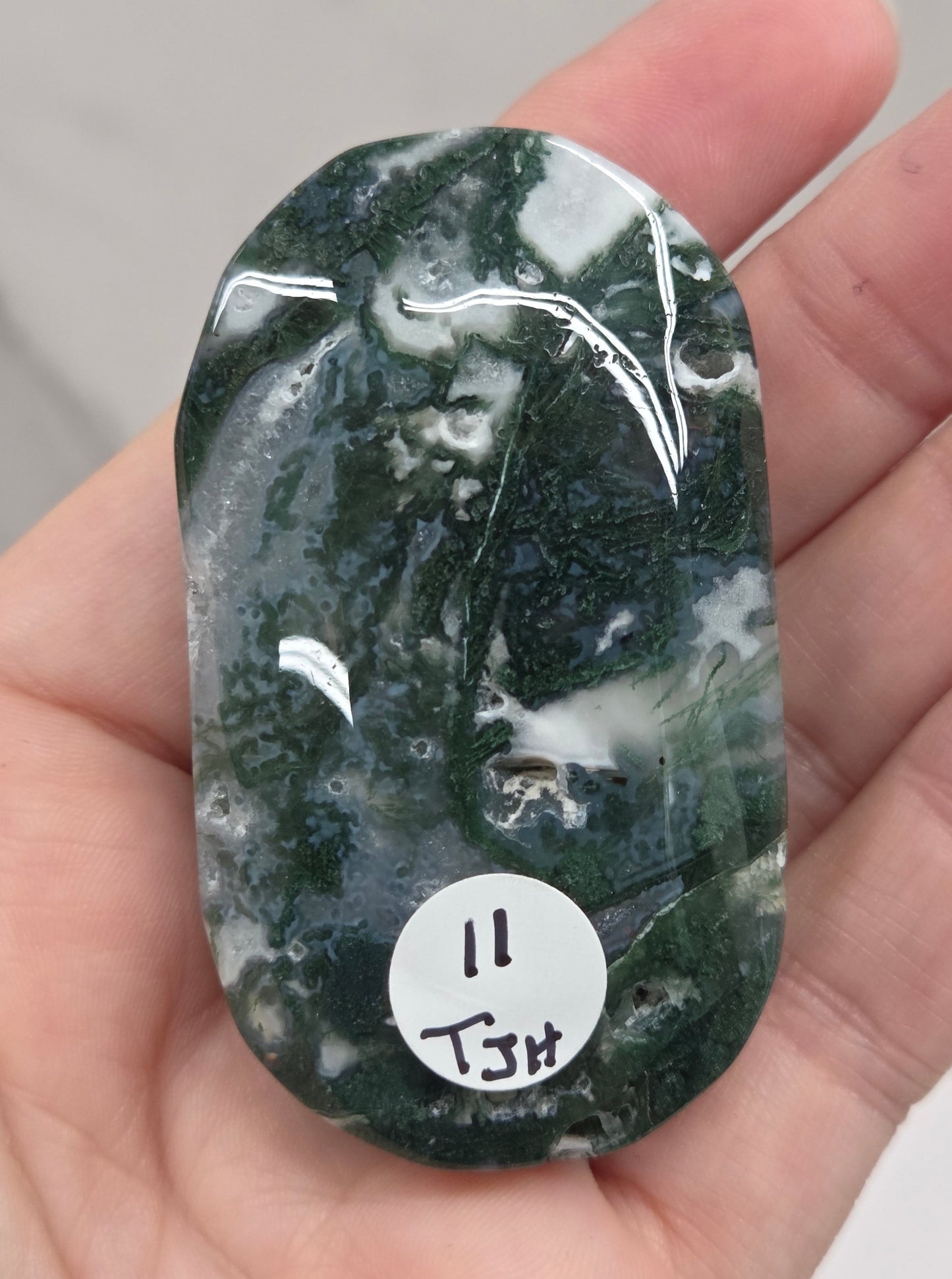 A hand holding a polished dark green and white marbled stone or jade-like oval piece, with a small white circular sticker on the right side.