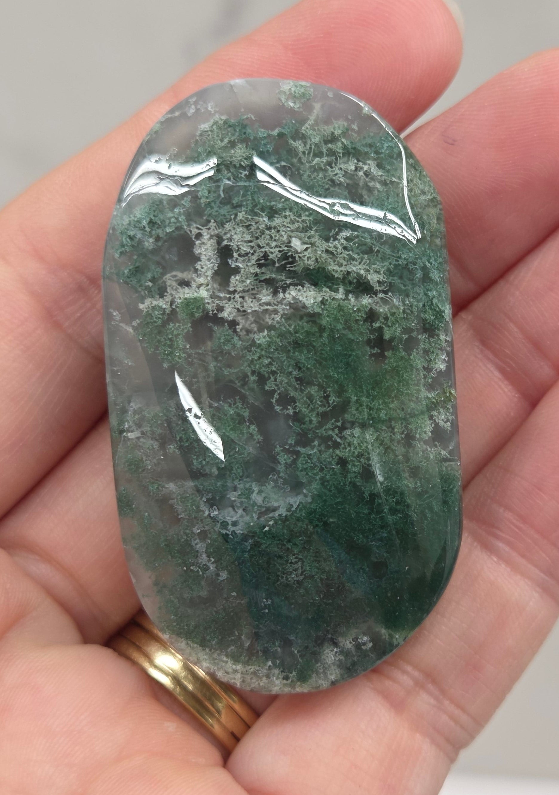 A hand holding a polished green stone with natural mottled patterns, showing its glossy, smooth texture.