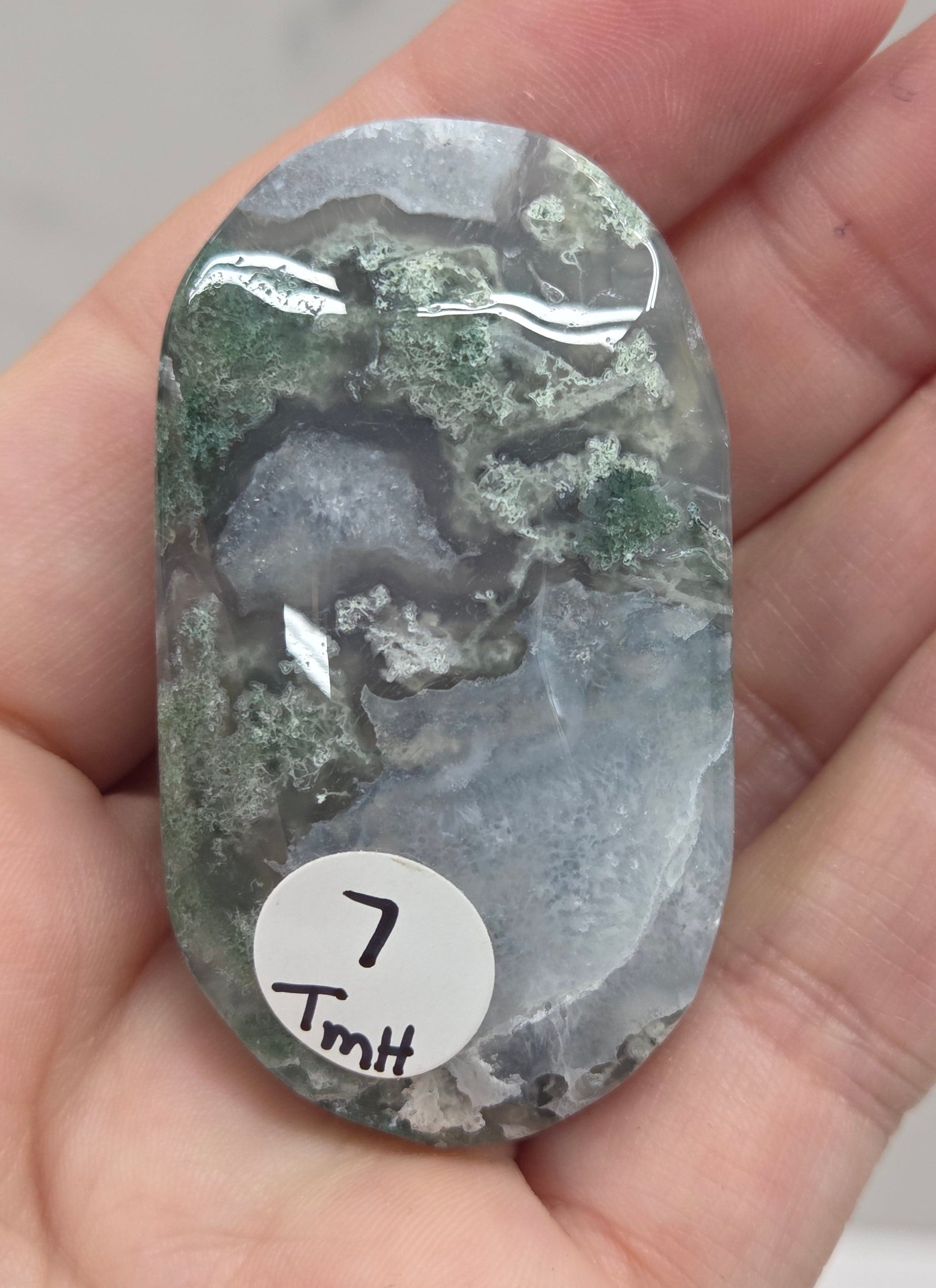 A hand holding a polished, rounded natural stone with green, gray, and white marbled patterns. A small white circular sticker with black characters is attached to the stone.