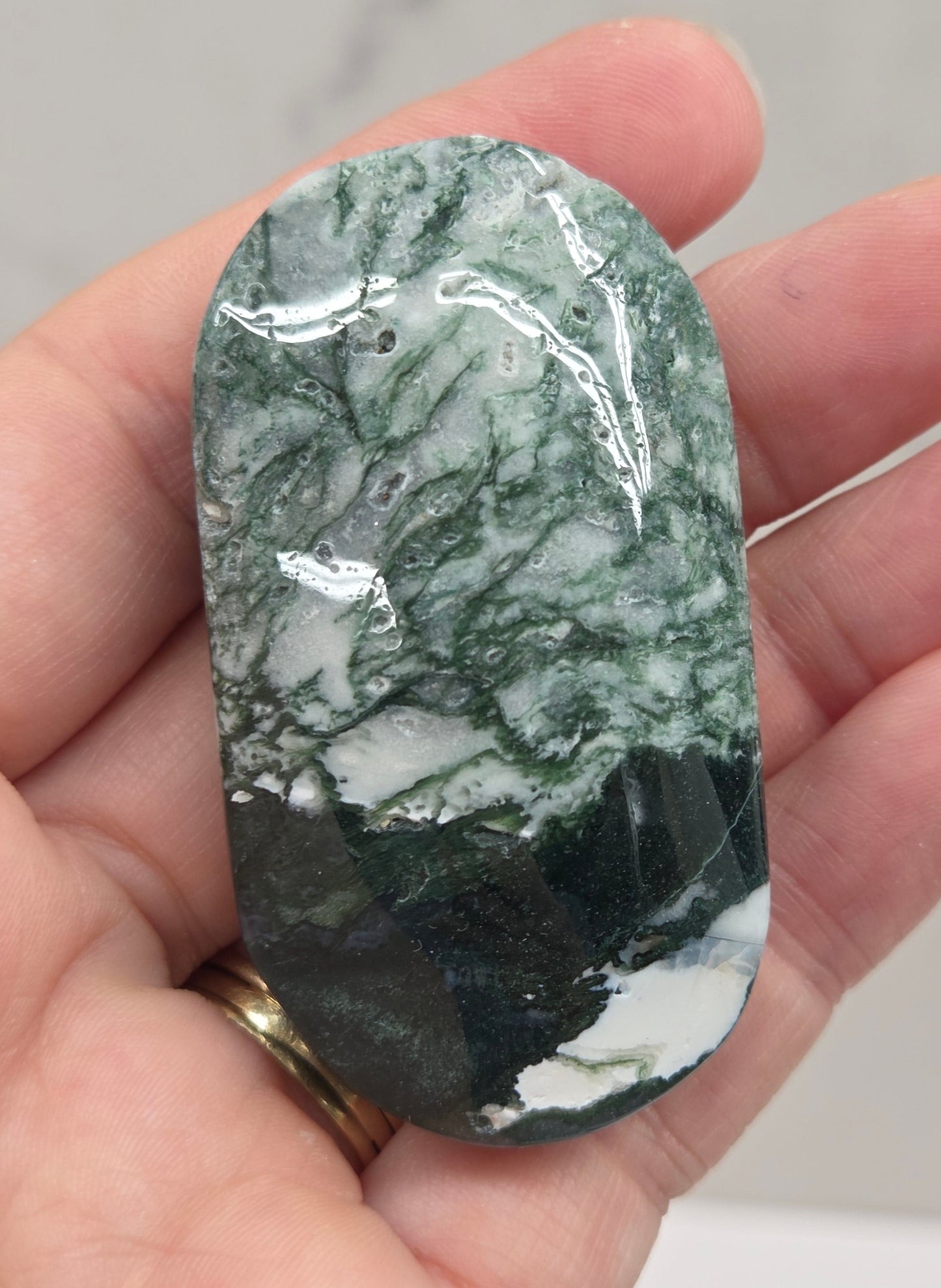 A polished green and white marbled stone pendant held in a person's hand, with a gold ring on one finger, against a light gray marble-patterned background.