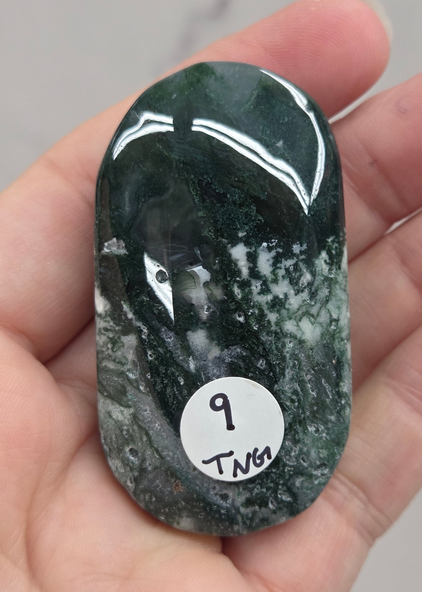 A polished dark green and white marbled stone slab held in a person's hand, with a small white sticker labeled '9 TMT' on the surface.