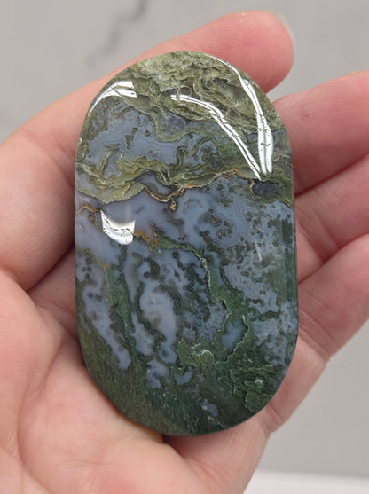 A polished green and gray marbled stone held in a person's hand, showing natural veining and a smooth rounded shape.