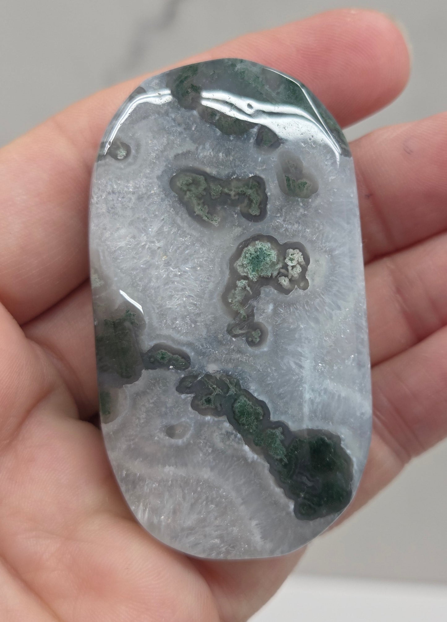 A polished stone or crystal slab with natural marbled patterns in gray, white, and dark green, held in a person's hand.