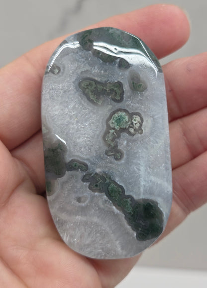 A polished stone or crystal slab with natural marbled patterns in gray, white, and dark green, held in a person's hand.