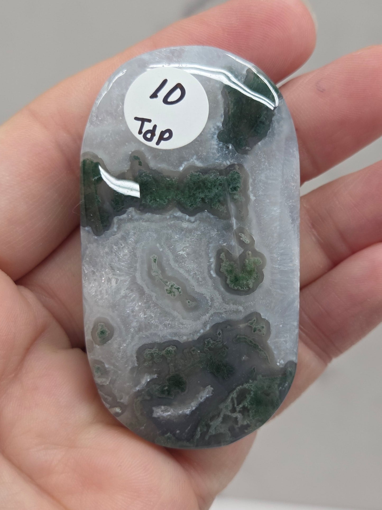 A hand holding a polished oval stone or chip with a white sticker that reads '10 Tap'. The stone has a light gray and white marbled pattern with dark green inclusions, photographed against a light gray marble-like background.