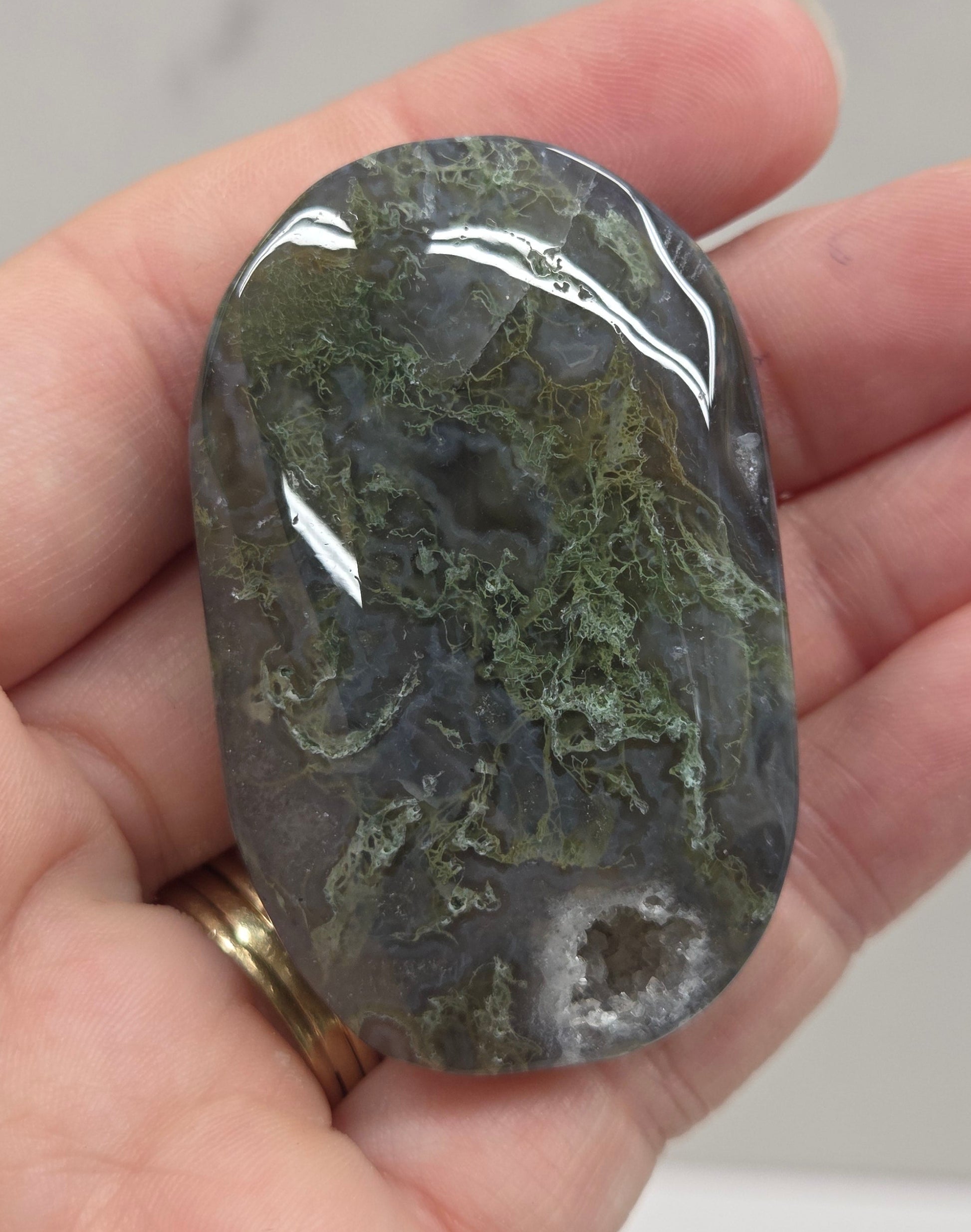 A polished dark gray and green stone held in a person's hand, with a gold ring visible on one finger against a light gray marble-like background.