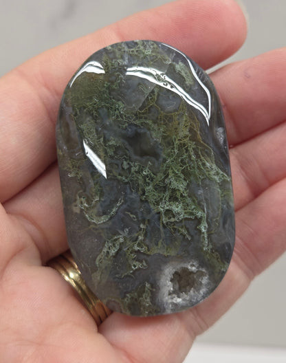 A polished dark gray and green stone held in a person's hand, with a gold ring visible on one finger against a light gray marble-like background.