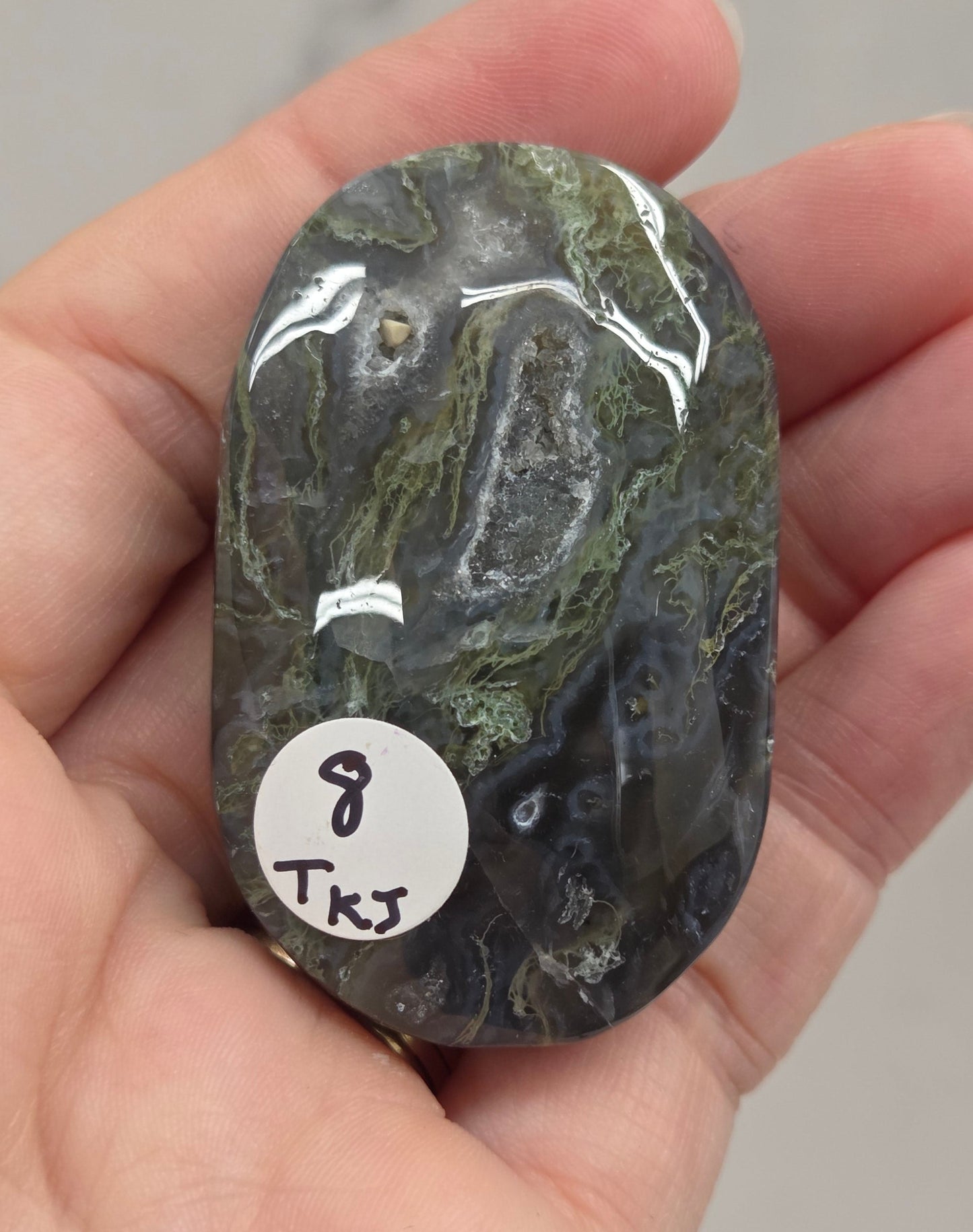 A hand holding a polished dark green and black marbled stone with a small white sticker labeled '8 TKS' on it, against a light gray marble-like background.
