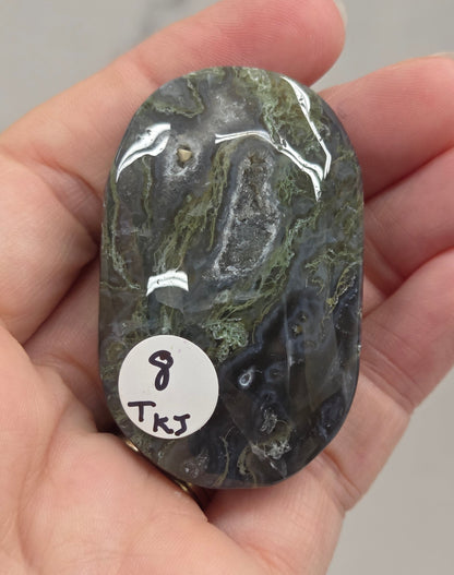 A hand holding a polished dark green and black marbled stone with a small white sticker labeled '8 TKS' on it, against a light gray marble-like background.