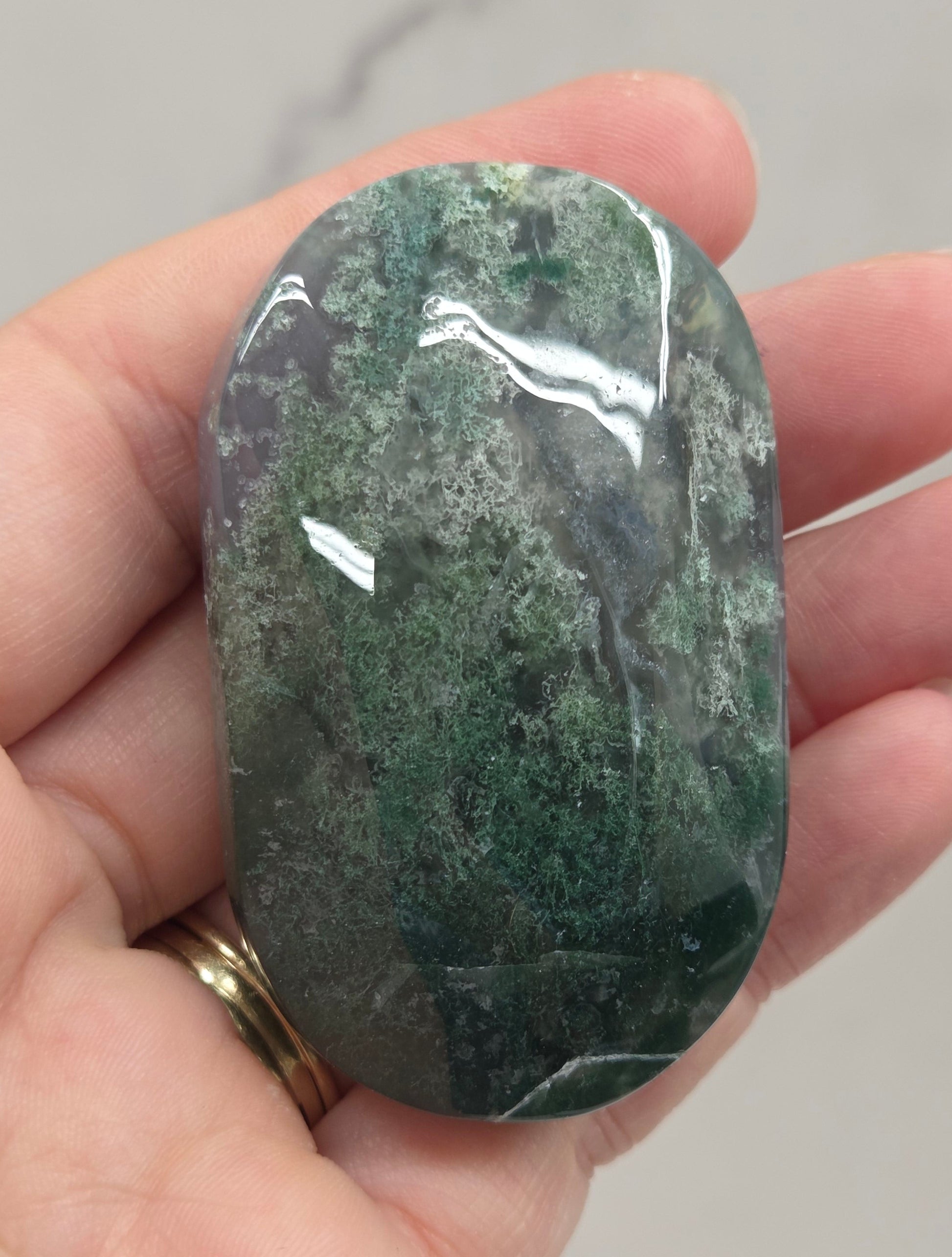 A polished green stone with natural mottled patterns, held in a person's hand. The stone has a glossy finish with light reflections, and the hand is positioned against a light gray marble-patterned background.